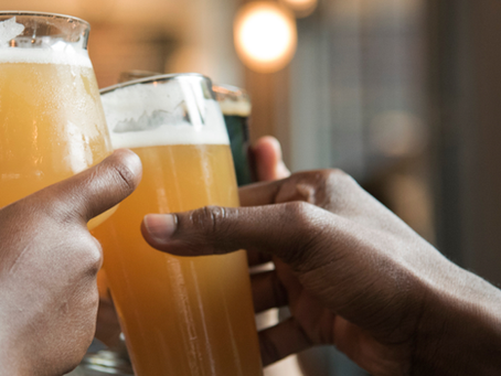 Mãos de pessoas brindando com copos de cerveja alaranjada em bar, com sorrisos desfocados ao fundo.