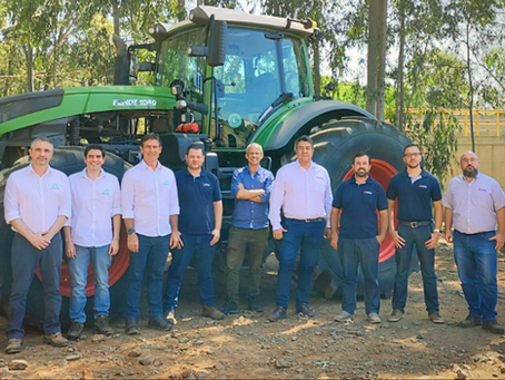 Grupo de 10 homens posa diante de trator verde em área arborizada, em clima de equipe.