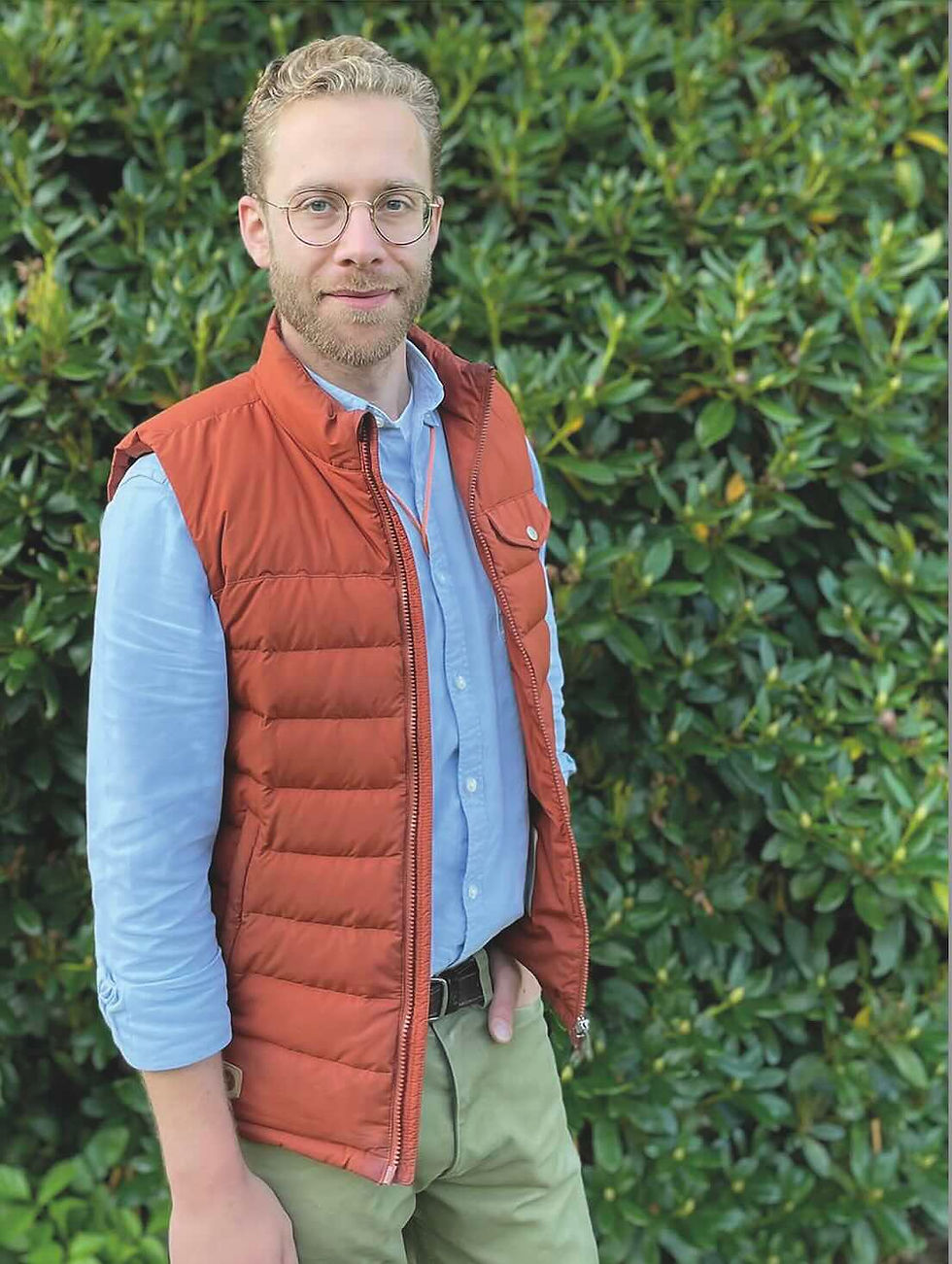 Dr. Johannes Gerst, bekleidet mit einer roten Weste und einem blauen Hemd, steht vor einem Hintergrund aus dichten grünen Sträuchern und lächelt leicht in die Kamera.