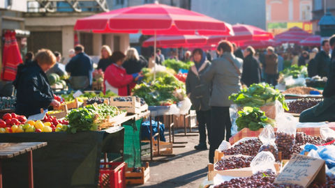 Marché en plein air animé, étals de fruits et légumes sous grands parasols rouges, clients et vendeurs au soleil.