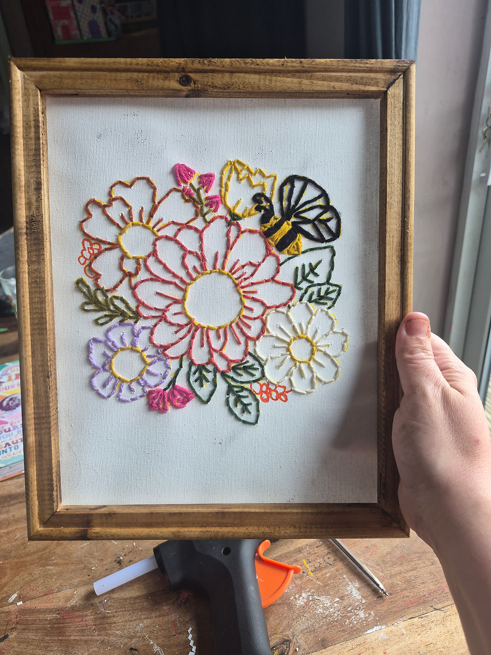 Hand holds framed embroidery of colorful flowers and a bee on white canvas, over a craft table.