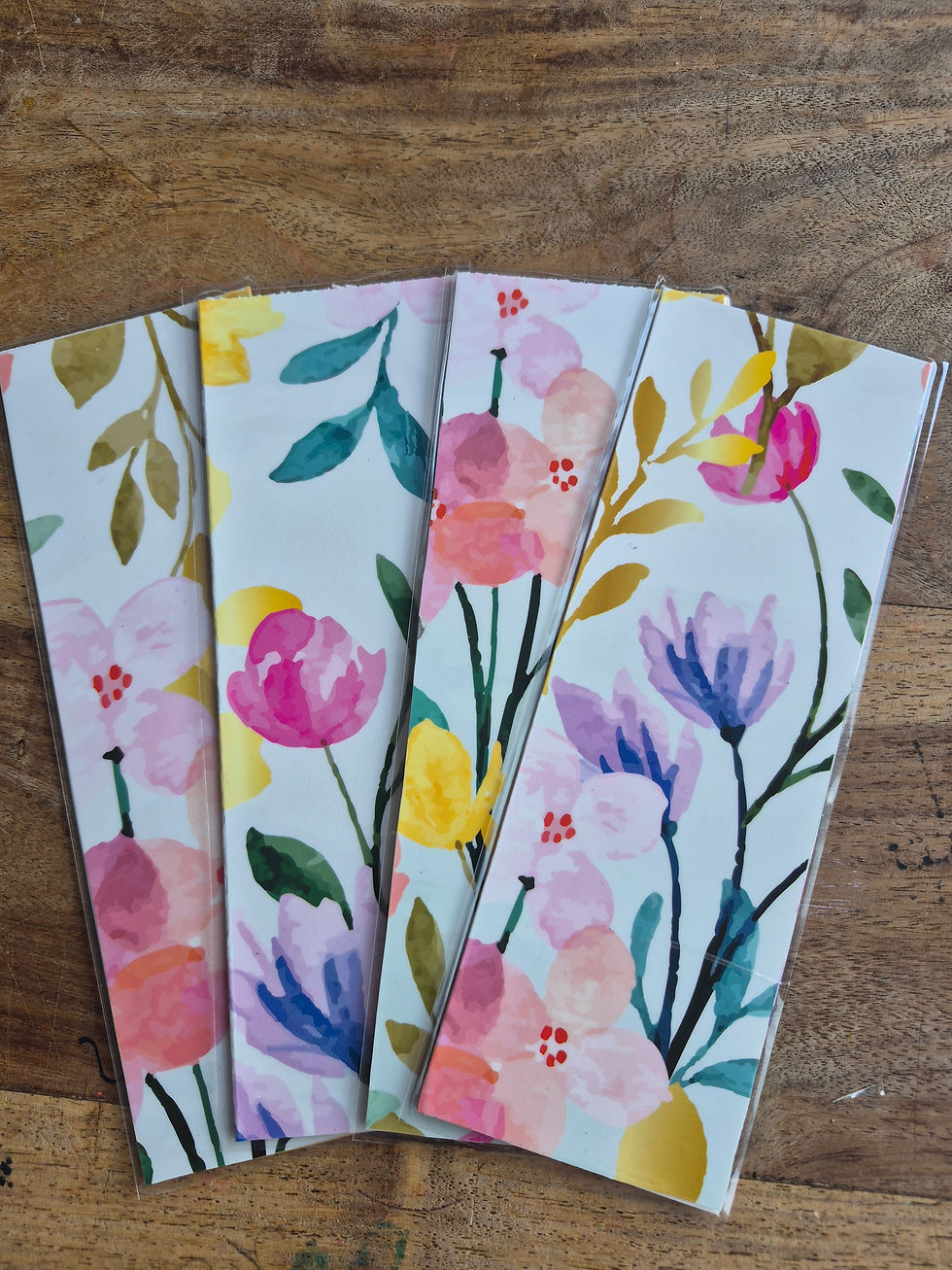 Four colorful floral bookmarks in clear sleeves fanned on a rustic wooden table, with pink, yellow, blue, and purple blossoms.