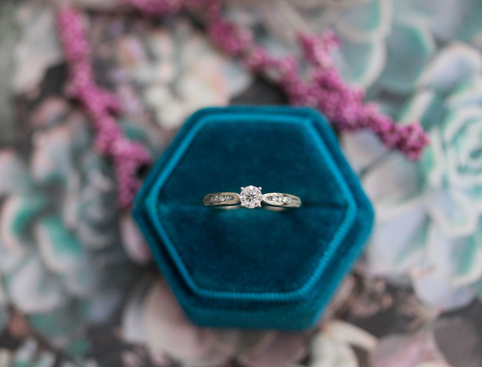 Diamond ring on teal velvet hexagonal box, placed on floral-patterned background with blurred lilac flowers, creating an elegant and romantic mood.