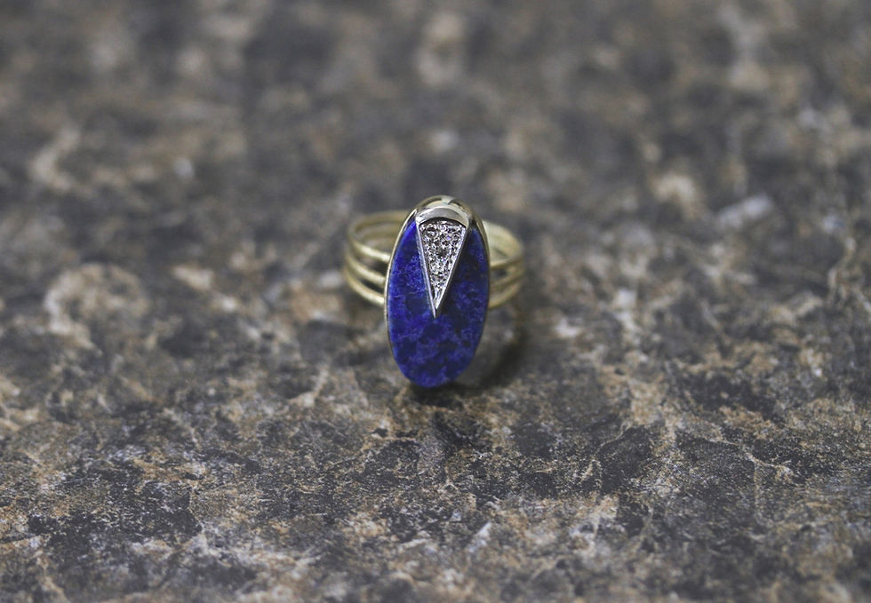 Gold ring with a blue oval stone featuring a triangular silver detail, set on a textured dark stone surface.
