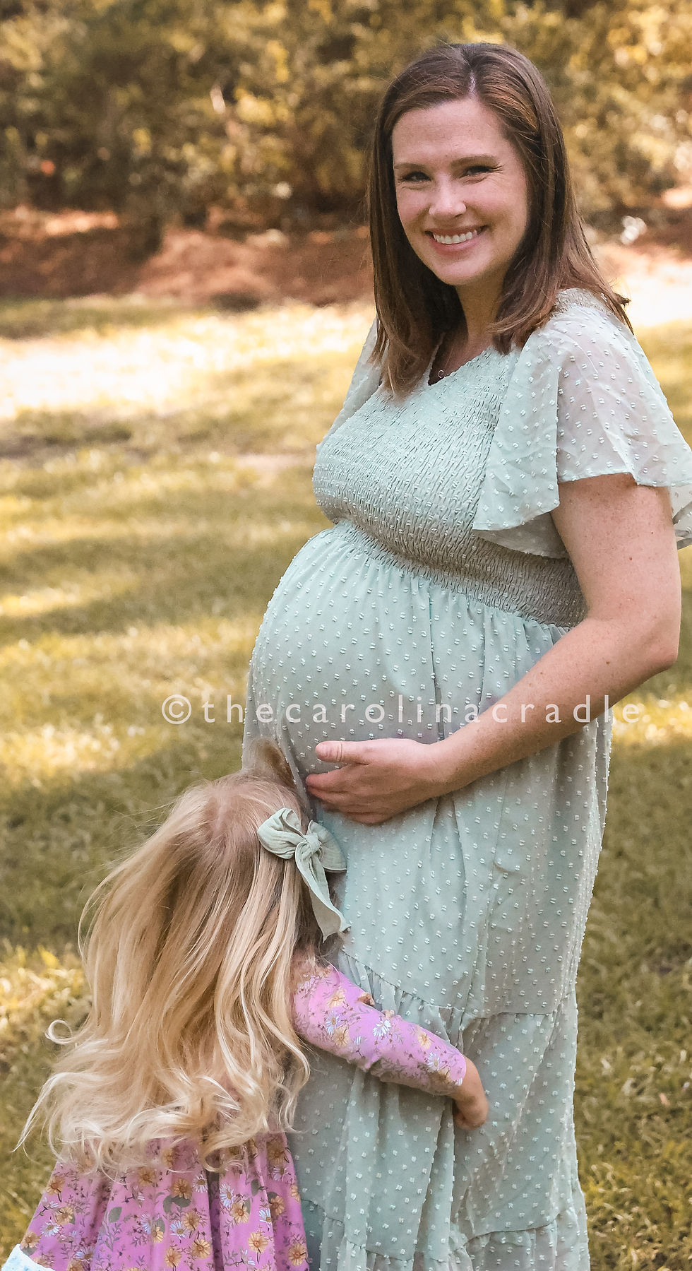 Pregnant woman in a light green dress smiles while a child with blonde hair in a pink floral dress hugs her belly. Sunny outdoor setting.