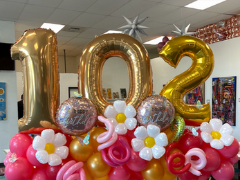 Balloon arrangement with gold '102', pink, white flowers, and 'Happy Birthday' balloons