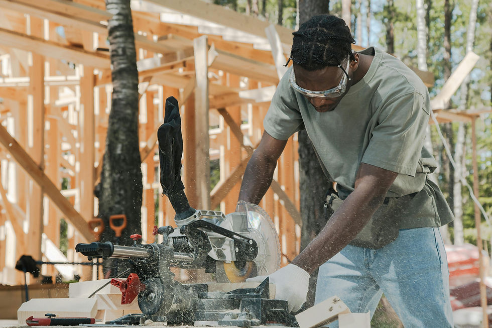 A contractor with dark skin and a grey shirt works a mitre saw in fromt of a new home construction with just the trusses