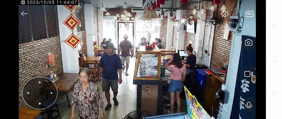 Casual photography of a lively, busy cafe interior with customers and staff mingling around a rustic wooden service counter. The atmosphere is bustling and casual with eclectic hanging light fixtures and brick accents.