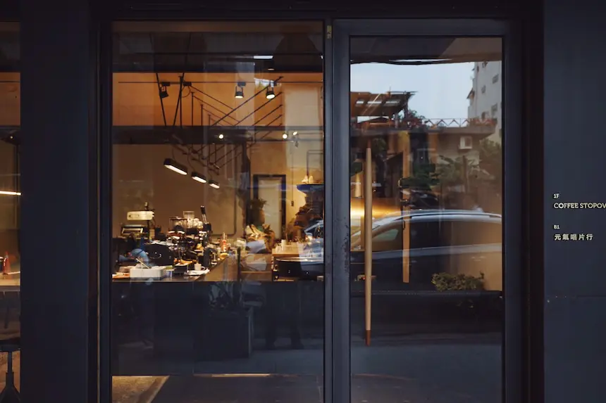 咖啡馆的玻璃门透出温暖的室内光线，内部可见咖啡机和器具，外面停着一辆车，门上有英文和中文字样。