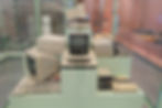 Vintage computers on display, with monitors and keyboards stacked in a glass case. Soft green and peach background in a museum setting.