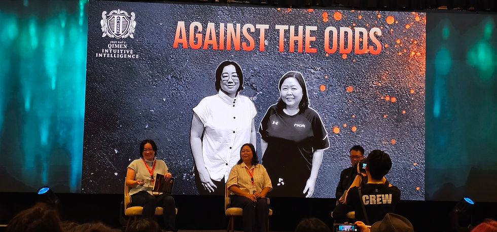 Two women sit on stage in front of a screen with "AGAINST THE ODDS" text. Background features a textured pattern and blue-green lighting.