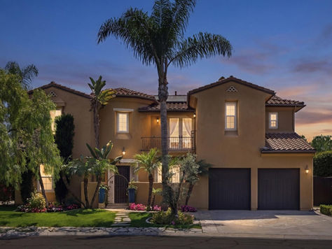 Twilight view of the luxury Mediterranean-style estate at 31491 Carril De Maderas in Rancho Madrina, San Juan Capistrano. The two-story home features a warm stucco exterior, arched windows, a private balcony with wrought-iron railings, and a two-car garage. The lush front landscaping includes mature palm trees, a manicured lawn, and a stone walkway leading to a gated courtyard entry under a vibrant sunset sky.