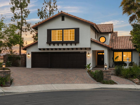 A beautifully updated Southern California home at 22231 Stillwater is captured at twilight on a quiet corner lot. The white stucco and terracotta tile architecture are framed by lush tropical landscaping and mature palm trees under a colorful sunset cloud formation. Warm light glows from the plantation shutters, highlighting the dark wood accents and double garage door. A meticulous brick paver driveway and stone retaining walls with lit lanterns enhance the welcoming, secure environment of this guard-gated Canyon Crest residence.