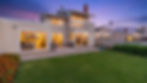 Captured against a stunning twilight sky, the rear exterior showcases the home's seamless blend of indoor and outdoor coastal living. Expansive sliding glass doors open the warmly lit interior revealing glimpses of the inviting bedroom and living spaces directly onto a spacious concrete patio, creating an effortless flow for evening relaxation.