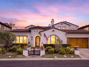 Front exterior of 31482 Paseo Campeon, a Spanish-style luxury home in guard-gated Rancho Madrina, San Juan Capistrano, at sunset — featuring a white stucco facade, red clay tile roof, arched wrought-iron front door, herringbone mahogany garage door, and mature landscaping with a wrought-iron entry gate.