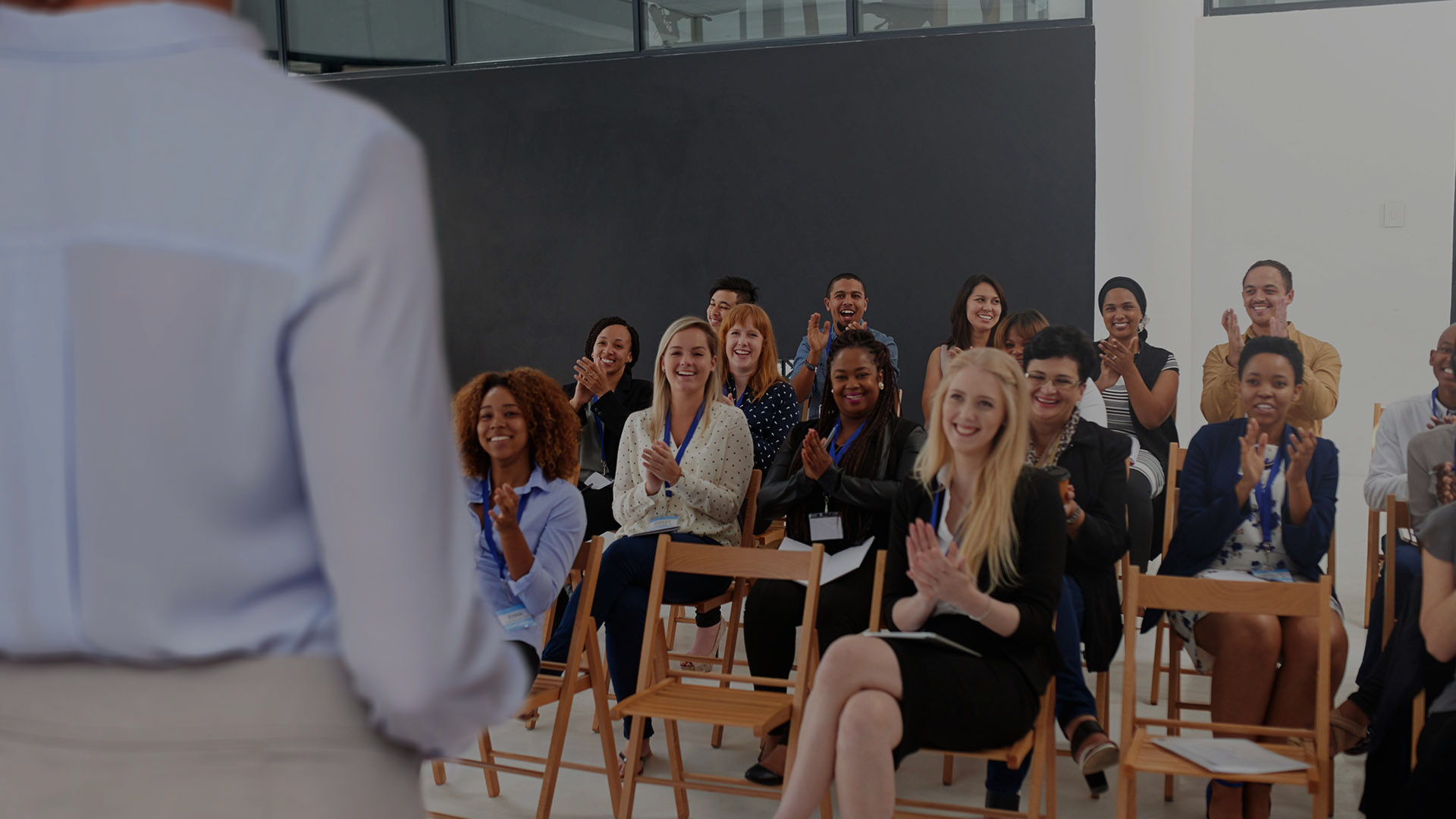A man speaks to a diverse group of colleagues gathered around laptops in a bright, modern office with large windows.