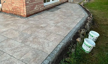 Neue graue Pflasterterrasse am Backsteinhaus, mit zwei Eimern im Gras.