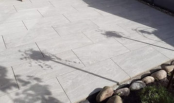Helle, gepflasterte Terrasse mit Schatten und Steinrand im Garten.