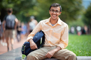 Bright outdoors portrait photo of a male Indian college student. He is wearing a peach-colored shirt and glasses, holding a blue backpack, and smiling.