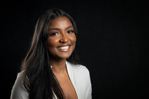 Photo portrait of a young black female college student. She has long black hair. She's wearing a white top and has a nice smile and the background is black.