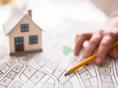 Main pointant sur un plan cadastral avec une maison miniature, représentant la vérification d'un terrain avant achat immobilier en Suisse