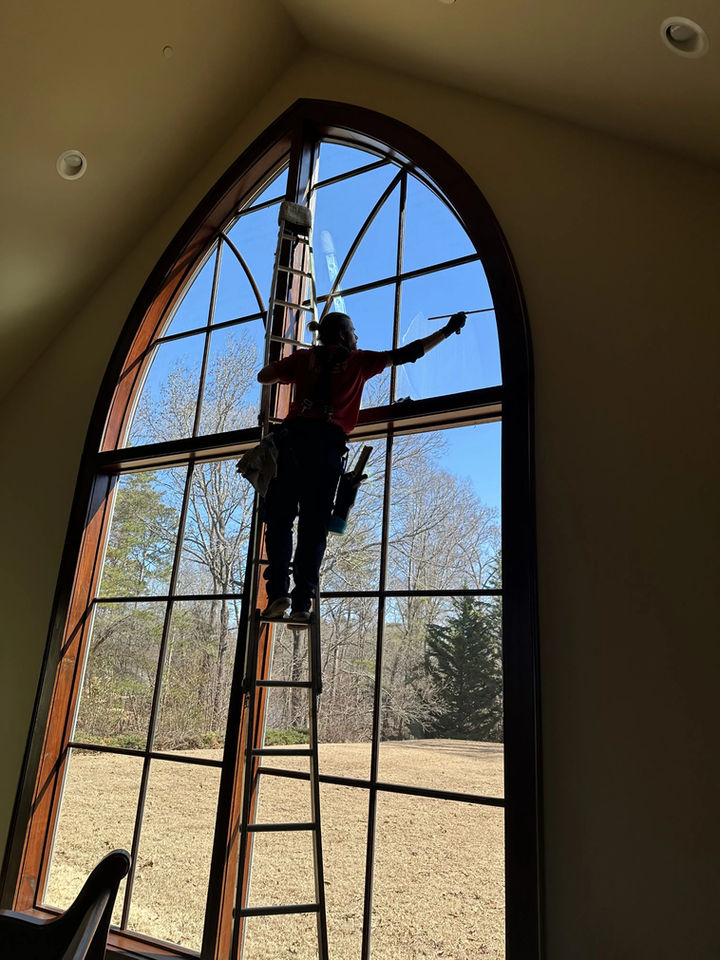 Worker on a tall ladder cleaning a large arched window inside a high-ceilinged room