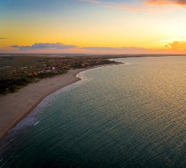 Vista aerea di una costa al tramonto, con una spiaggia che si curva dolcemente lungo le acque tranquille e una cittadina che si estende lungo la riva. Il cielo sfumato di arancione e rosa si riflette sull'acqua, creando una scena pacifica.