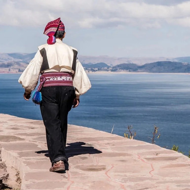 Un abitante di Taquile cammina su una strada di pietra panoramica con vista sul Lago Titicaca, che regala un riflesso di un cielo sereno, in una scena pacifica che invita a esplorare le ricchezze culturali durante un viaggio in Perù.