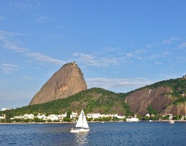 Una barca a vela bianca naviga sulle acque blu davanti al famoso Pão de Açúcar, una montagna a forma di panettone a Rio de Janeiro, sotto un cielo limpido e azzurro.