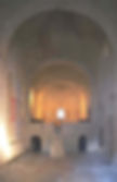 The austere interior of an ancient church in Acqualagna, featuring bare stone walls with arches and a simple altar at the end of a central nave, illuminated by soft light, creating an atmosphere of historic reverence.