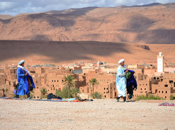 Due uomini in abiti tradizionali marocchini camminano davanti a una città storica con montagne sullo sfondo.