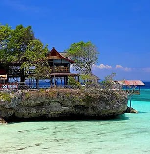 Un isolotto roccioso con una casa su palafitte circondata da acqua turchese cristallina, illustra il pittoresco paesaggio tropicale dell'Indonesia, perfetto per una fuga serena