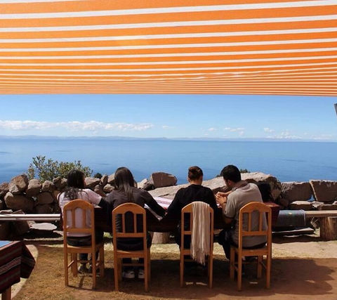 Sotto un vivace tendone a righe, una famiglia condivide un momento di unione ammirando l'immenso Lago Titicaca, durante il loro viaggio in Perù, una scena che incapsula la tranquillità e il legame affettivo, con l'infinità dell'acqua che si estende all'orizzonte