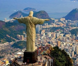 La statua iconica del Cristo Redentore si erge sopra la città di Rio de Janeiro con le braccia aperte, simboleggiando la benvenuta. In lontananza, si possono vedere le montagne e le isole al largo della costa, con la città vivace disseminata di edifici ai suoi piedi.