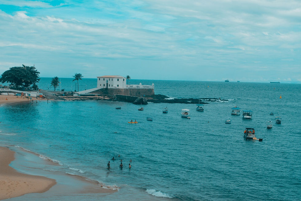 Una tranquilla scena costiera che mostra una spiaggia sabbiosa con alcune persone e barche ormeggiate nell'acqua azzurra. Una fortezza storica con mura bianche si affaccia sul mare, con palme che ondeggiano leggermente. Il cielo è nuvoloso, conferendo all'atmosfera un tono calmo e sognante.