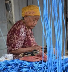 Una donna anziana indonesiana, con un tradizionale bandana gialla e abito marrone, è seduta e intenta a intrecciare fili blu vivaci, mostrando l'arte antica del tessuto a mano, uno scorcio di vita quotidiana e di artigianato tradizionale.