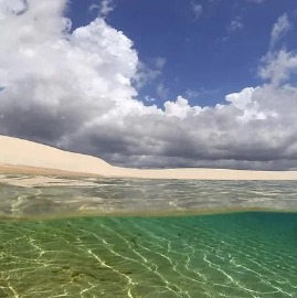 Una laguna cristallina situata accanto a dune di sabbia sotto un cielo drammatico con nuvole dense e luminose che suggeriscono un contrasto dinamico tra calma e tempesta.