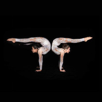 Two acrobats in white costumes performing a mirrored pose on a black background. 