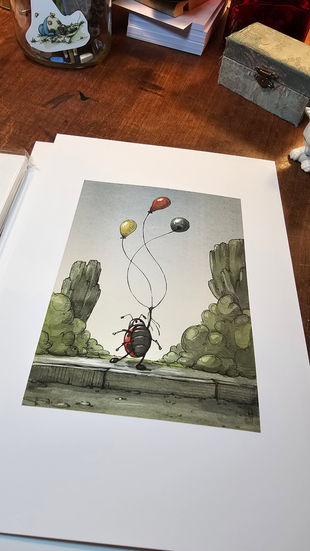 A photograph of a printed illustration lying on a wooden table. Illustration shows a watercolor painting of a ladybug holding three balloons the reach for the sky. She is standing on a gray pavement with green vegetation on either side and gray sky in the background. Balloons are colored yellow, red and blue. 