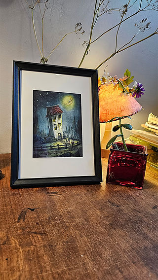 A photograph of artwork printed on white paper, in a black picture frame, standing on a wooden desk with a small plant in a red vase on the right. The artwork shows a mixed media illustration of a white house with a red roof placed with a dark blue background. The house is “looking” in the direction of a yellow moon that has a smiling face. Two people are walking in front of the houses. 