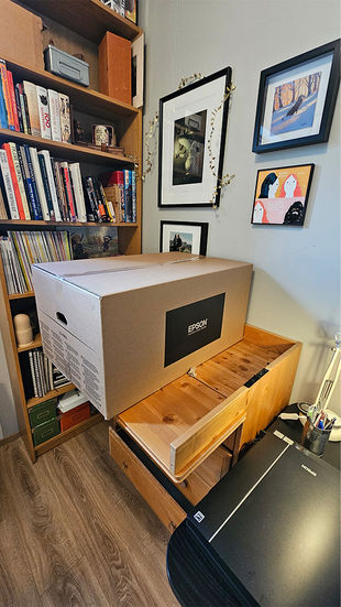 Photograph, from inside a small room, of a large cardboard box on top of a wooden drawer. The box is marked Epson. A bookshelf behind the drawer and pictures on the gray walls.