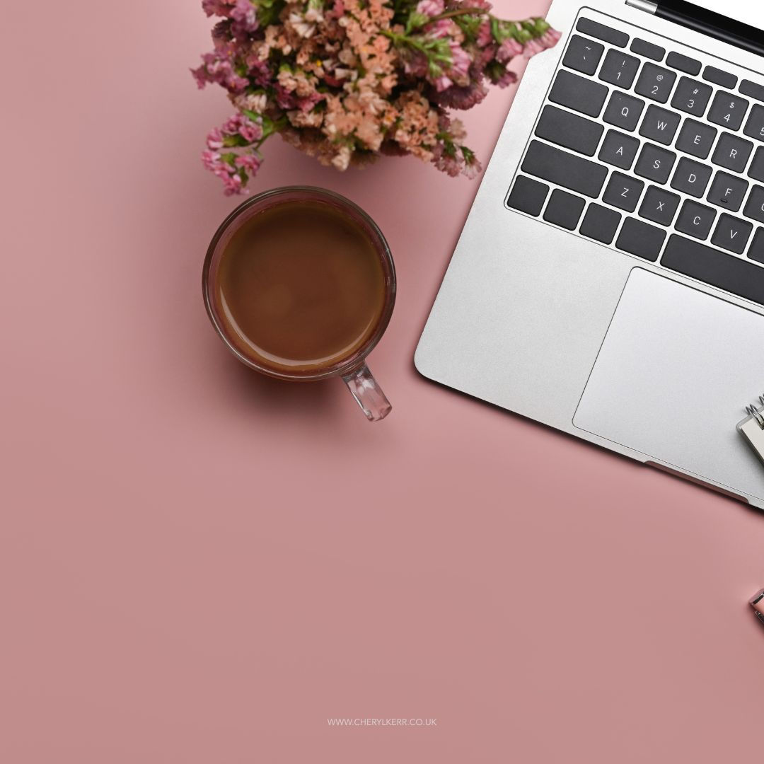 A rose gold macbook open on a table with a white mug and saucer next to it, in a mainly white room.
