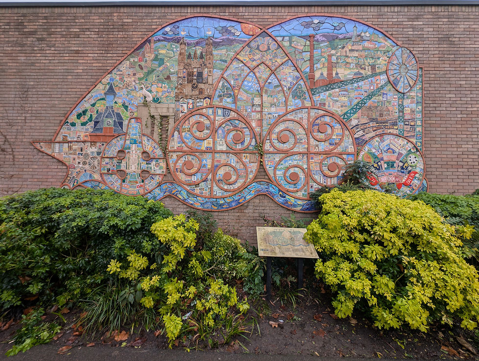 Colorful mural mosaic on a brick wall depicting urban scenes with swirls. Green shrubs in foreground, plaque with text visible. Vibrant and detailed.