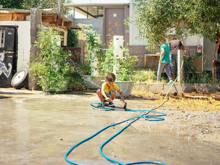 A boy joyfully plays with a garden hose, creating a spray of water in a sunlit backyard, surrounded by lush plants and rustic structures.