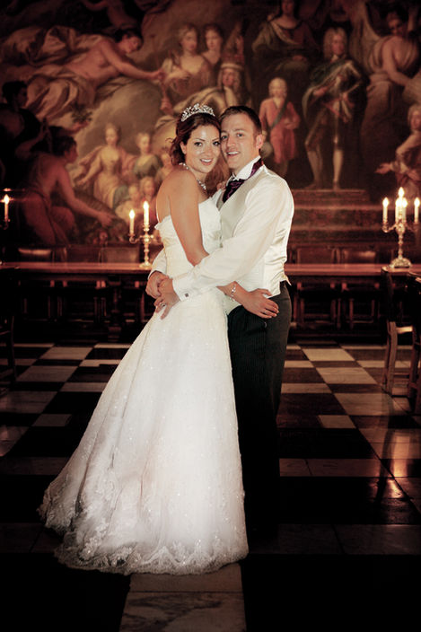 Bride and groom standing arm in arm in the candlelit Painted Hall at the Old Royal Naval College in Greenwich, surrounded by dramatic baroque murals and checkered marble flooring.