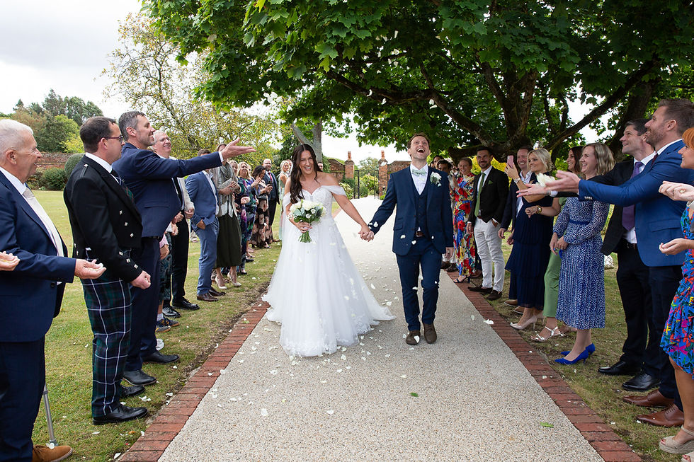 Just married at Gaynes Park, David and Sara walk through a shower of confetti surrounded by smiling guests under the trees of the Walled Garden