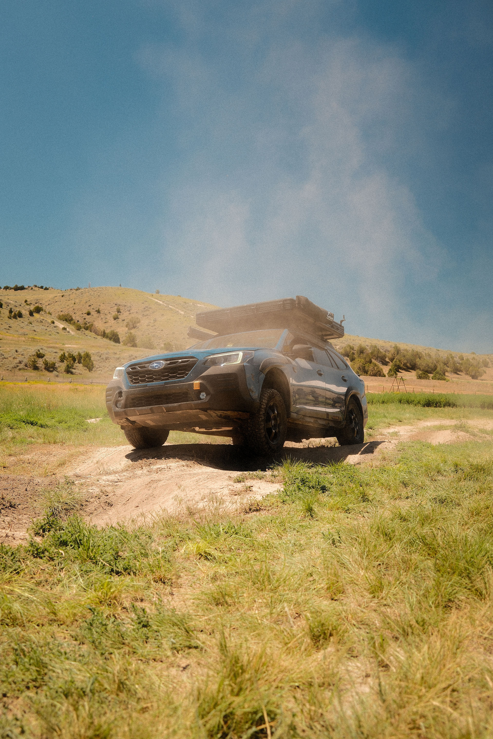 Blue Subaru on a dirt and grass trail with a rooftop tent on it. Mountains and blue sky in the background.
