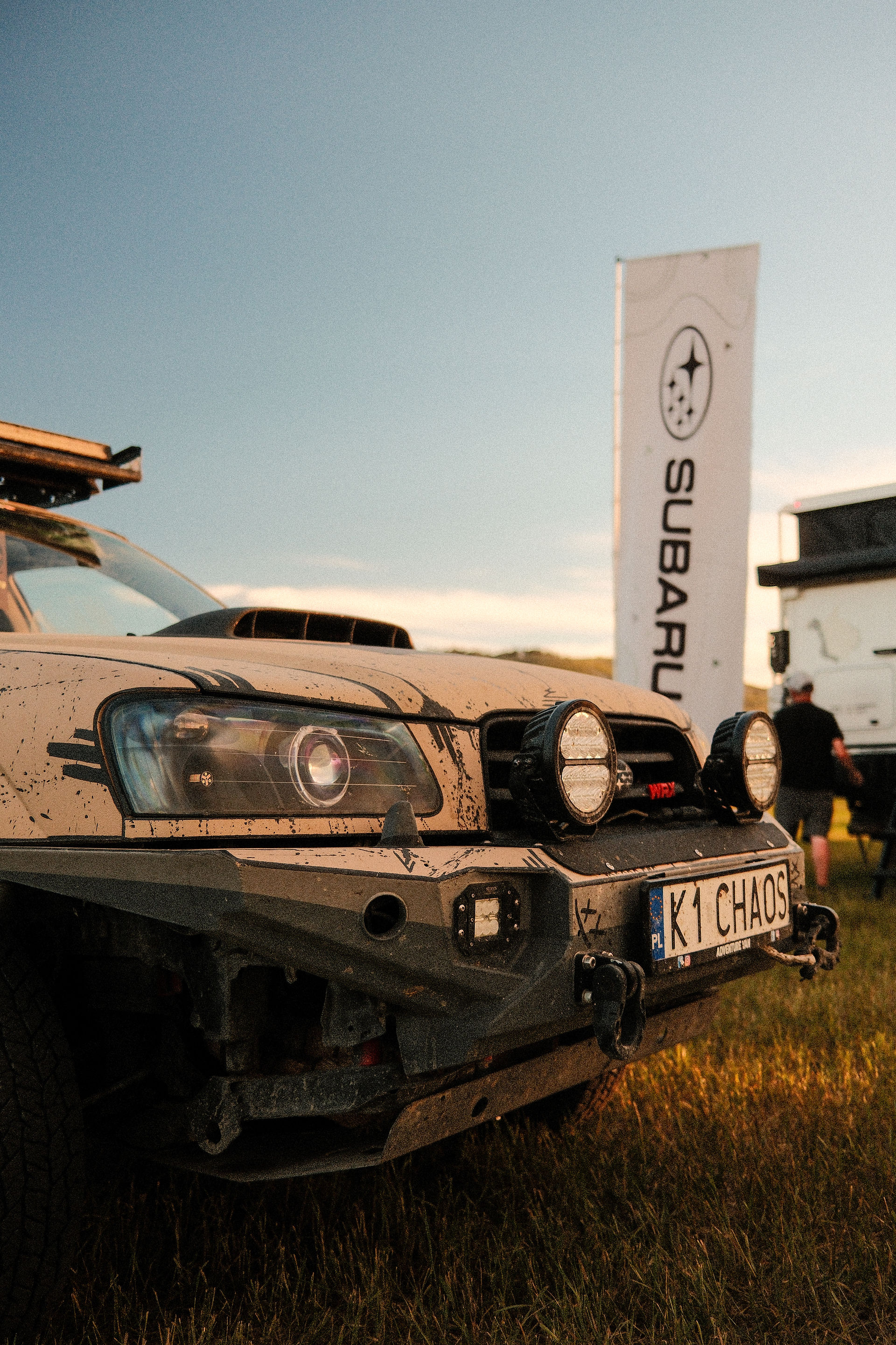 front 3/4 view of white subaru parked on grass. The car has an off-road bumper and lights. In the background is a white vertical subaru banner.