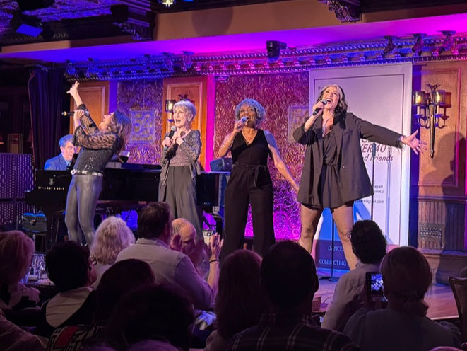 Four women from the original Chicago cast perform a vocal number at 54 Below, standing in front of the piano, backed by rich purple lighting and the venue’s ornate gold detailing.