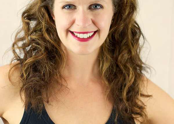 Professional headshot of Tara Memi smiling at the camera with long curly brown hair, wearing a black sleeveless top against a light neutral background.